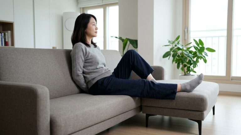 허리디스크 방사통 관련 사진, 밝은 한국 아파트 거실, 40대 한국 여성이 낮은 소파에 앉아 한쪽 다리를 앞으로 편안하게 뻗고 휴식하는 넓은 옆모습